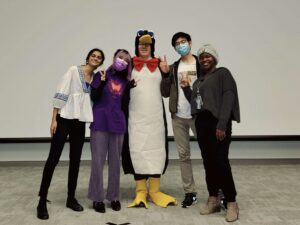 Donald Ainslie in penguin costume surrounded by four of his students, three presenting as women, one as a man, all of different ethnic backgrounds