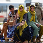 A group of young Afghans sitting on their belongings at the Kaburl airport, with a plane and military personnel in the background.