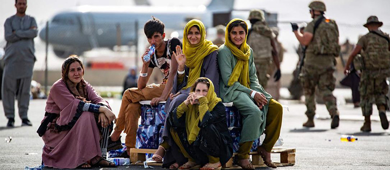 A group of young Afghans sitting on their belongings at the Kaburl airport, with a plane and military personnel in the background.
