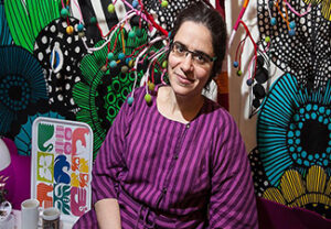 A bespectacled Agnes Callard dressed in a purple-and-black-striped dress sitting in front of a colorful wall of patterened artworks