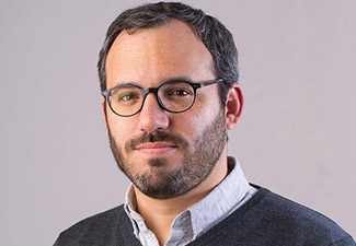 a headshot of a man with round glasses, a collared shirt, and a close-cropped beard.. He is smiling slightly.