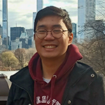 Head shot of a smiling Alex de Guzman against a backdrop of river and cityscape. Alex is Sotheast Asian, has short black hair, and is wearing glasses, a burgundy U of T hoodie, and a black parka over top.