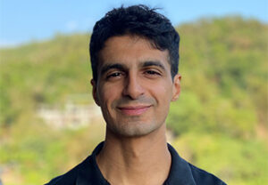 A clean-shaven man with short black hair wearing a dark polo shirt standing against a background of a grassy hill.