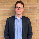 Upper body shot of Andrew Lavigne, a white man with short, light brown hair wearing glasses, a white T-shirt under a blue button-down shirt and dark gray blazer. He is standing against a brick wall and smiling.