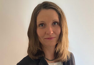 A middle-aged white woman with shoulder-length dark blonde hair wearing business attire and a simple bead necklace.