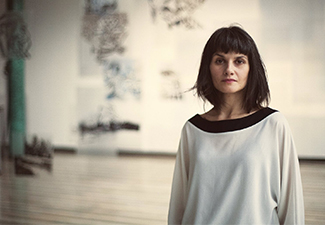 Bara Kolenc, a middle-aged woman with shoulder-length dark hair wearing a loose, long-sleeved white top with a broad black neckline, standing to the right in a wide open studio space with blurred paintings and sculptures in the background.
