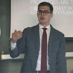 Head shot of Benjamin Brewer lecturing in front of a projection. He has short, dark hair, glasses, and is wearing a gray suit, white shirt, and red tie.