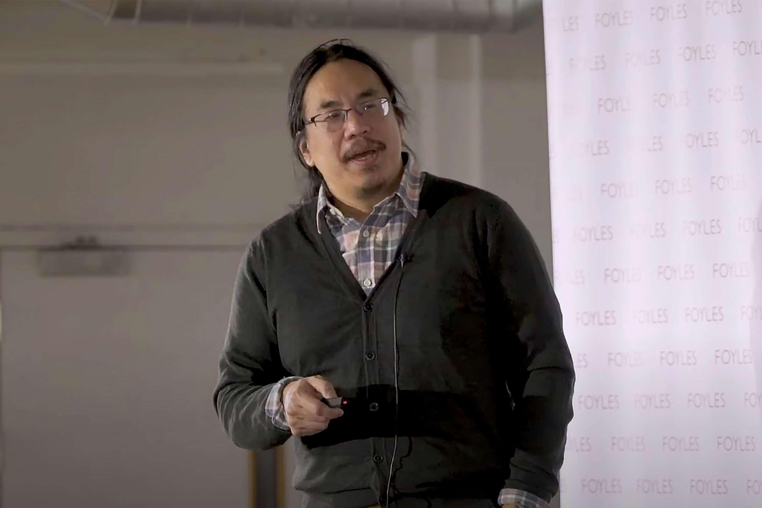 An East Asian man with long hair tied in a loose ponytail and wearing glasses and a brown sweater in the midst of giving a presentation, pointer in hand.