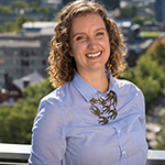 Head shot of a smiling Caitlin Hamblin-Yule wearing a blue blouse and necklace of bronze hands and standing against a Toronto cityscape from above. She is a white woman with long, curly blonde hair.