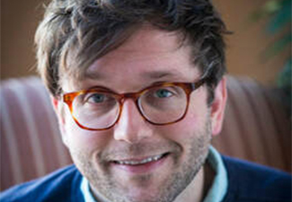 Close-up of Chris Howard, a white man with red glasses and casually messy short hair wearing a blue shrt and pullover, grinning into the camera.