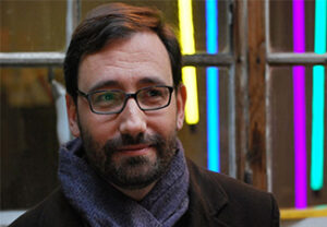 Head shot of Claude Romano, wearing a winter coat, scarf, and glasses, standing in front of a window with brightly colored light tubes showing in the background