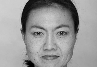 Black-and-white close crop of Curie Virág, an unsmiling, middle-aged East Asian woman with her long black hair tied in a low ponytail