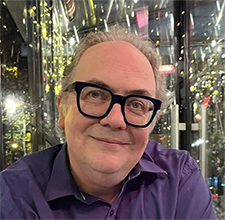 Man with glasses and purple shirt outside with lights in evening.