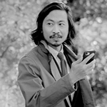 Black-and-white image of David Suarez, and Southeast Asian man with shoulder-length hair and a goatee, wearing a suit and trnechcoat and holding a cell phone looking to the left in front of shrubs