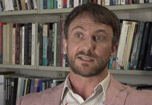 A white man with a short beard sitting mid-conversation in front of a bookshelf wearing a collared shirt under a light blazer.