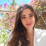 Head shot of Eirini Martsoukaki, a woman with long brown hair wearing a white summer top sitting against a backdrop of a pink flowering bush.