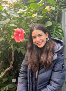 Elif standing next to a green plant with large red flower.