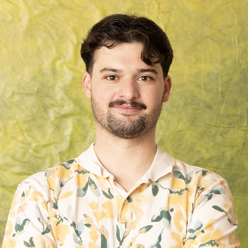 Head shot of Evan Drapeau, a young white man with short brown hair and a mustache and scruffy short beard wearing a polo shirt covered in yellow flowers and standing in front of an abstract green background, smiling slightly.