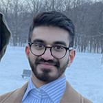 Head shot of a smiling Faisal Bhabha against a winter landscape. Faisal has short, well-coiffed black hair, a short beard, wears glasses, and is wearing a white-and-blue striped dress shirt under a camel-colored jacket.