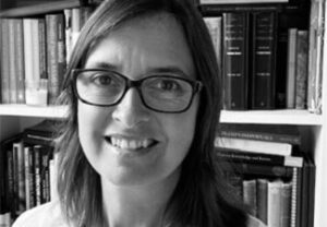 Black-and-white close-up of a smiling and bespectacled Fiona Leigh in front of a bookshelf