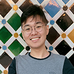 Gerald Teng, a young East Asian man with short black hair and glasses wearing a gray and black long-sleeved T-shirt, standing in front of a colorful mosaic background and smiling broadly into the camera.