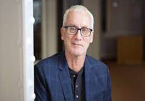 Head shot of Huw Price, a white man with white hair wearing glasses, a black shirt, and a blue blazer, standing against the perspectival backdrop of an office, leaning against a door