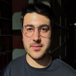 Head shot of Jason Singer, a white man with dark, short hair, glasses, a some beard scruff, wearing a charcoal sweatshirt and standing in front of a shaded wall.