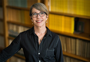 A short-haired, bespectacled Jessica Wilson wearing a black shirt standing smiling in front of a bookshelf full of yellow-spined journal covers.