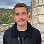 Head shot of Jogn Tzatzanis, a white, blond man with a mustache, standing wearing a black casual jacket an T-shirt in front of a brick structure and a field blooming with wild flowers