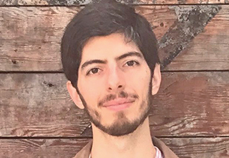 Head shot of a young man with short brown hair and a short beard, leaning casually against a barn door wearing a button-down shirt and jacket.