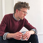 Image of Jonny Cottrell, a young man with short blond hair, glasses, and wearing a burgundy sweater with a long-sleeved shirt underneath, sitting looking away from the camera as though in thought, holding a mug.