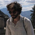 Head shot of Joseph Gerbasi, a man with dark, middle-length, curly hair and a beard, wearing a white shirt and sun glasses against a mountain backdrop.