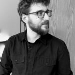 Black-and-white shot of Joshua Brecka, a white man with a beard and glasses wearing a black shirt standing by a door