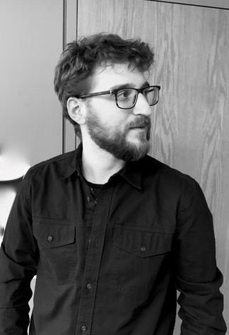 Black-and-white shot of Joshua Brecka, a white man with a beard and glasses wearing a black shirt standing by a door