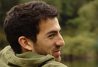 Head shot of Justin Bledin wearing an olive-gree hoodie, his back turned to the camera but his faced turned toward it. He has short, dark hair and a stubble beard and the image is set against a blurry forest background.