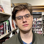 Head shot of Kacper Mykietyn, a white man with short, dirty blond hair, wearing glasses and an olive green bomber jacket, standing in front of library shelves