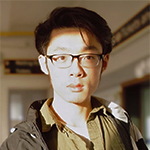 Head shot of Kangyu Wang, a younf East Asian man with short black hair and glasses wearing a kaki button-down shirt and olive jacket standing inside an interior with high windows