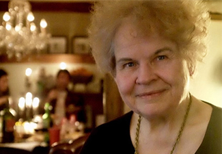 Head shot of an elderly Kathryn P. Morgan to the right of the frame, smiling into the camera, with a festive dinner scene in the blurred background.