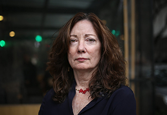 An oder Latina woman with long, dark hair wearing a black top and red necklace standing in front of a restaurant window with a serious face facing the camera.