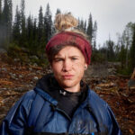 Outside head shot of Lis McMillan, a white woman with a top knot wearing a red headband and blue anorak against a forest background