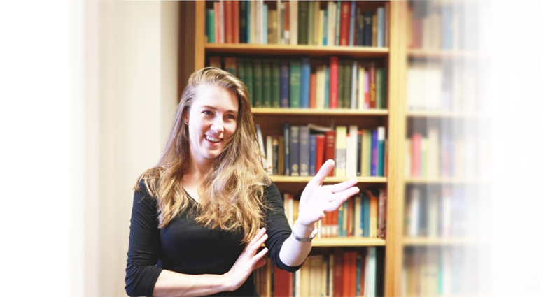 Lu-Vada Dunford gesturing toward the viewer standing in front of a bookcase