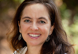 A headshot of a woman with shoulder-length brown hair . She is smiling towards the camera. She wears silver dangling earrings