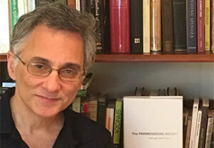Head shot of Michael Della Rocca, a middle-aged man with short gray hair and glasses wearing a black shirt, standing smiling beside a book shelf.