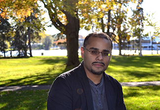 Head shot of Mohammed Rustom, who has short dark hair, a short beard, and glasses, sitting in a beautifully lit park with a lake and trees.