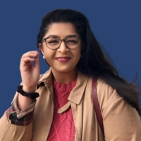 Nipa Chauhan. who has long black hair and glasses, stands smiling in front of a U of T blue background wearing a salmon-colored top under a beige jacket
