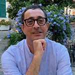 Head shot pf Pasquale Porro, a middle-aged man with short dark hair, glasses, and a blue-and-white striped shirt sitting in front of a blooming blue bush, his chin on his hand