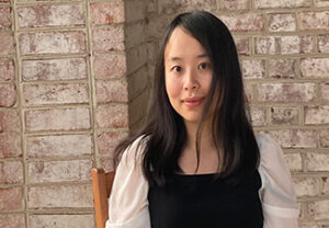 A young East Asian woman with long black hair sitting on a wooden chair in front of a brick wall, wearing a white, puffed-sleeve blouse under a black dress.