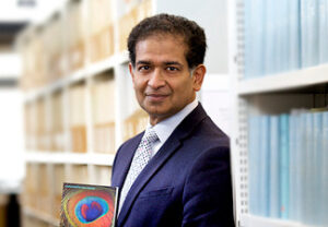 Chakravarthi Ram-Prasad, an Indian man wearing a blue suit, stands at a 3/4 angle towards the viewer. Behind him are shelves of orange and blue books.