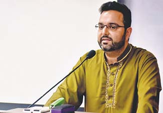 Sajjad Rizvi, bearded, bespectacled, and wearing a mustard yellow shirt or shalwar kameez, standing at a lectern
