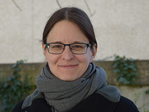 A woman with long, dark blond hair tied together in the back and wearing glasses, a winter jacket, and a green scarf standing outside with a house and bits of bare branches visible, smiling